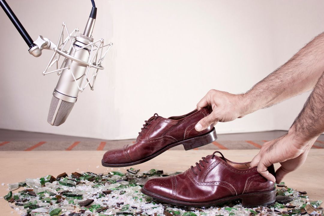 About Us 267 A foley artist is manipulating a pair of brown leather men's shoes over a surface of broken glass and stones. A microphone hovers above the shoes and glass.