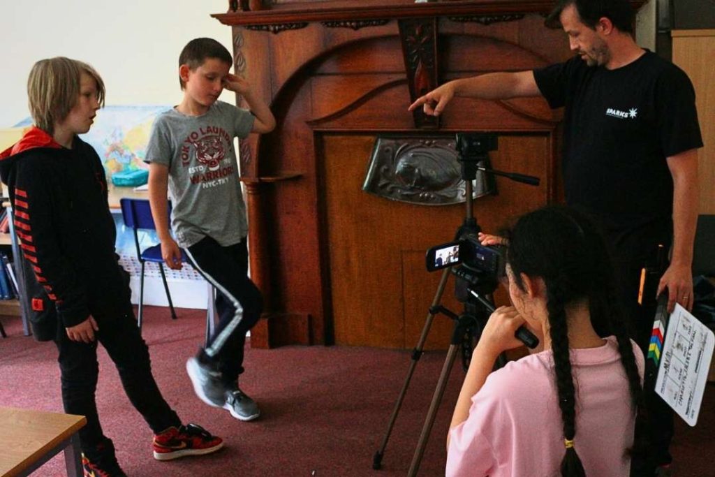 Alyn, Head of Sparks FIlm School Cardiff West, frames up a shot with a group of three young filmmakers. Alyn is pointing and giving directions, two students stand in front of a camera on a tripod, and one stands behind, directing. 