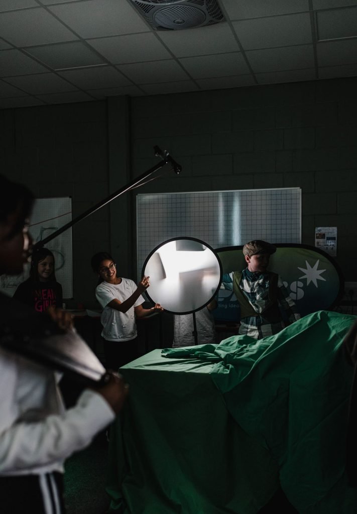 Sparks Film School Courses - Film and Media Courses for Ages 5-18. A young film crew are gathered on set in a dark room. There is backlighting and a boom pole in the air. 